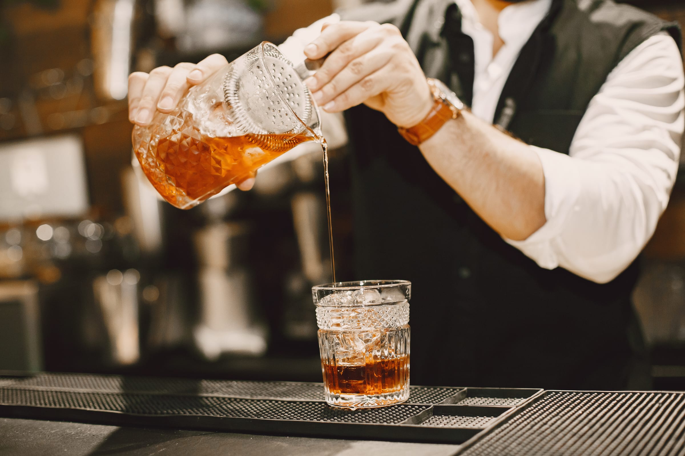 Bartender in a white shirt and vest pouring a stirred amber cocktail from a cut-glass mixing pitcher into a rocks glass over ice